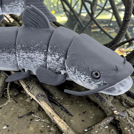 Tidal 3D Printed articulated grey catfish toy with realistic mottled texture and barbels resting on muddy ground.